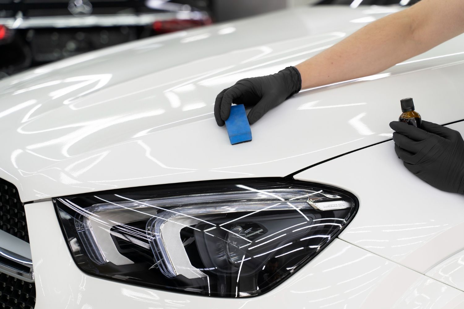 Gloved hands apply protective film to a white car hood and headlight in a workshop
