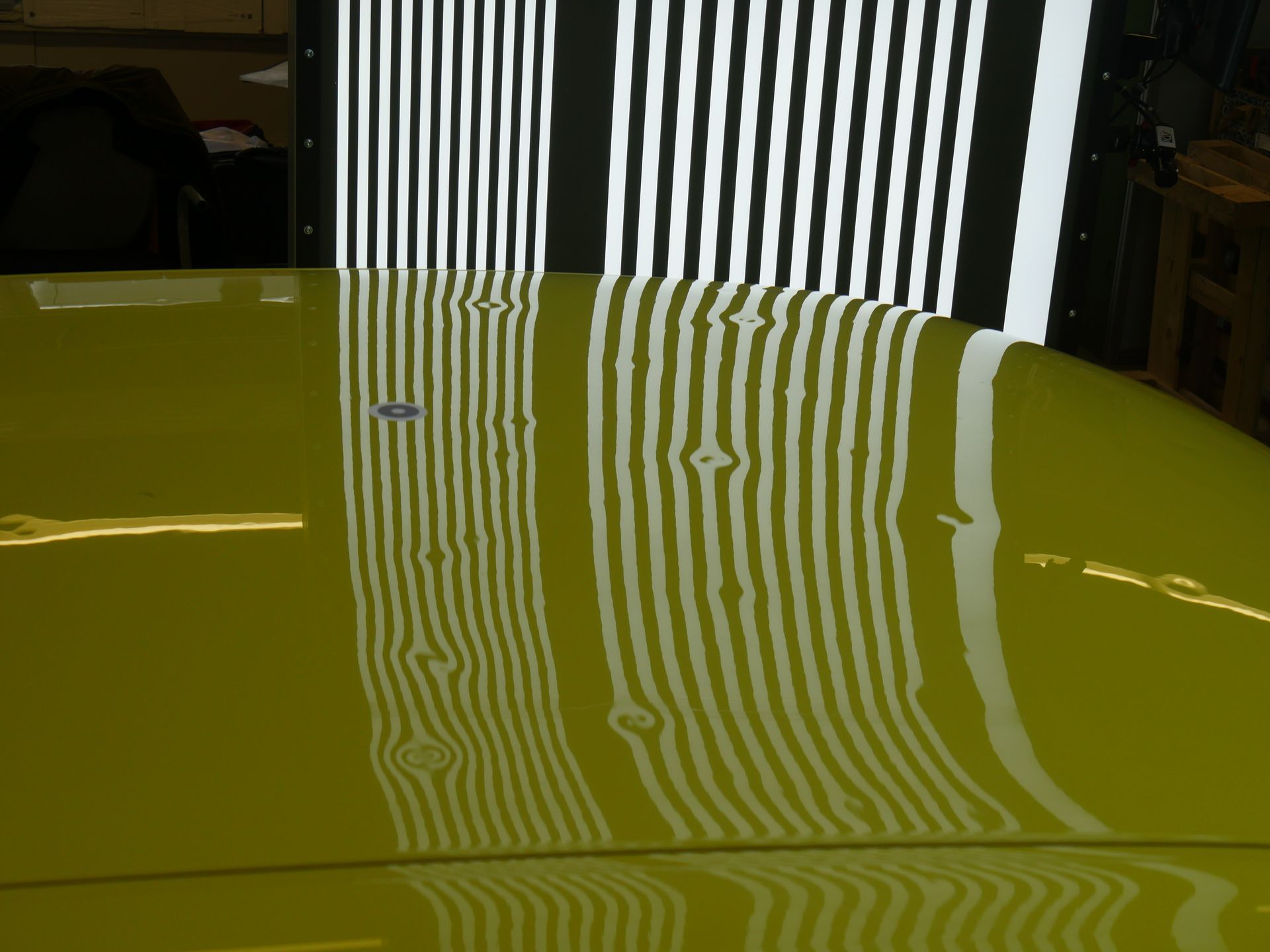 A yellow car hood being inspected for dents using a reflected grid board light.