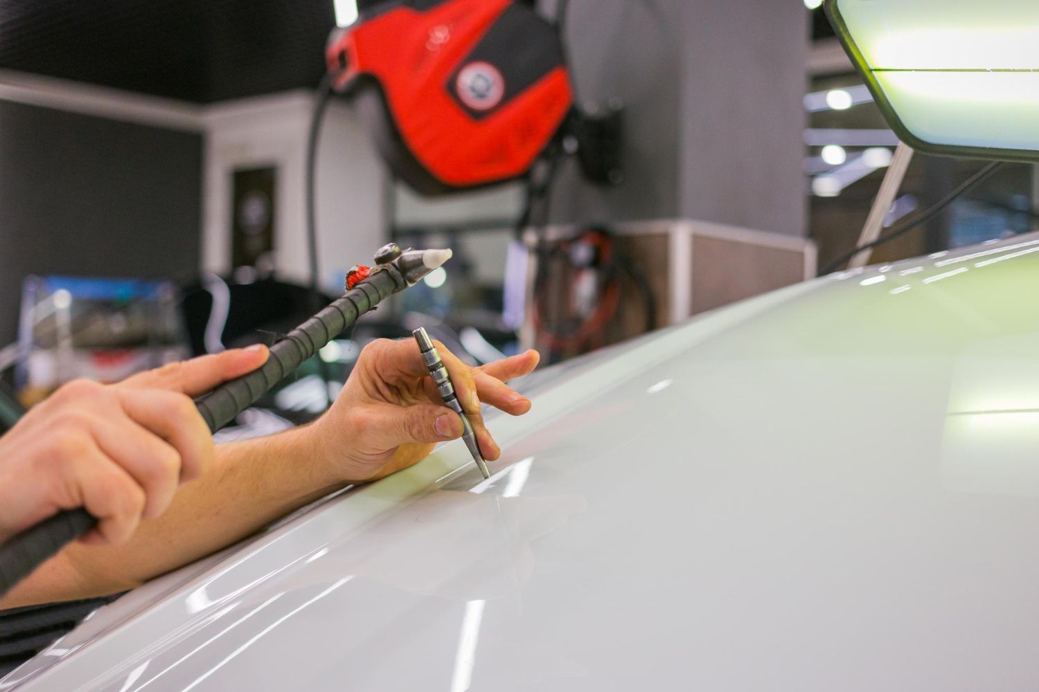 A technician uses a specialized tool to perform paintless dent repair on the white hood of a car in a workshop.
