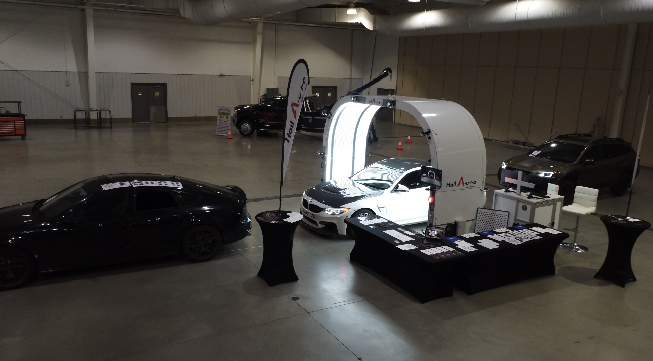 A brightly lit, arched photo booth displays a car at an indoor event, with black vehicles parked on either side.