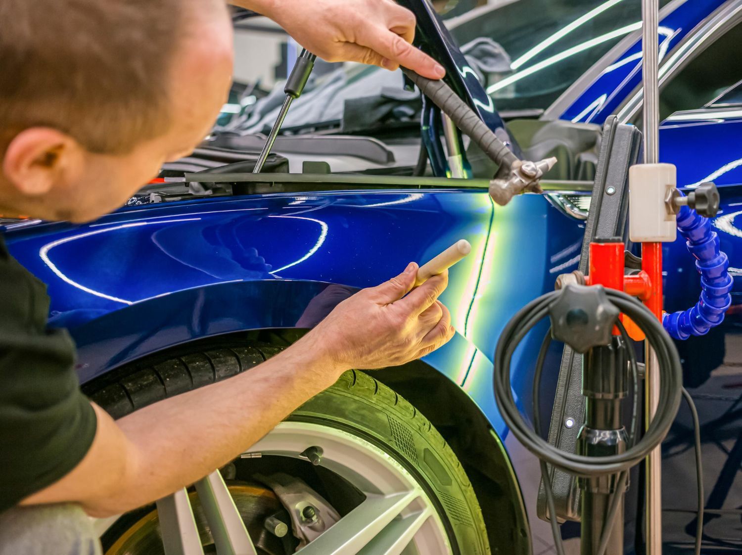 Technician welding or repairing the blue car's front fender in an auto shop
