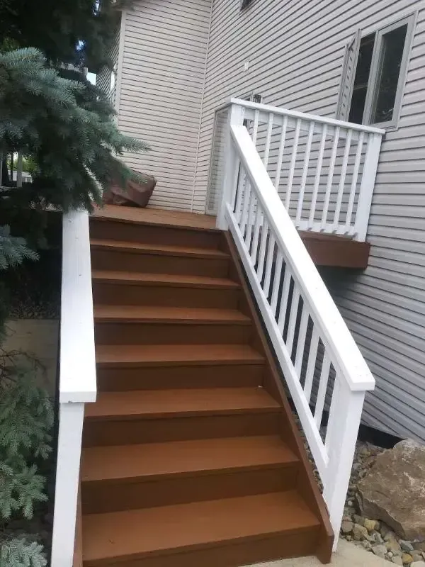 Brown wooden stairs with white railings leading up to a house porch beside brick siding.