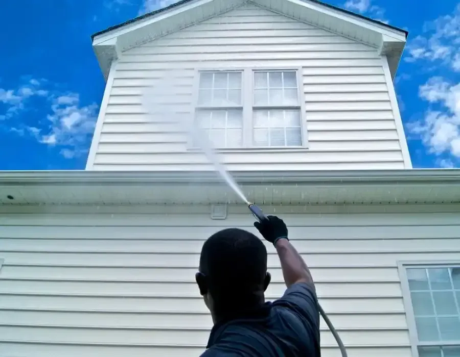 Person pressure-washing a white house exterior under a blue sky