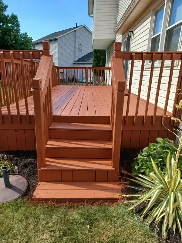 Red wooden backyard deck with steps and railing beside a house, surrounded by grass and plants