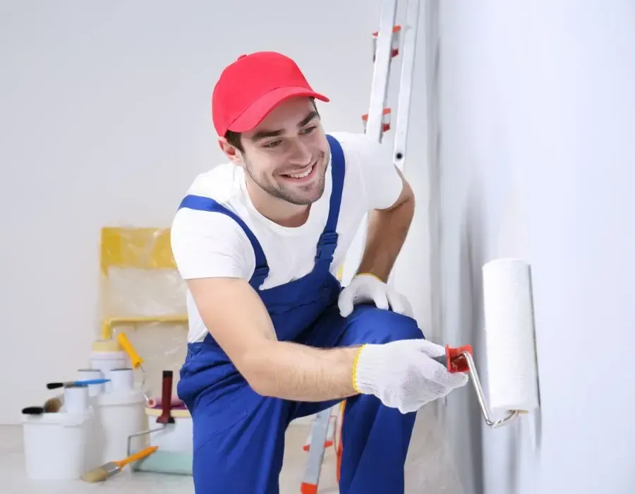 Painter in blue overalls and red cap painting a white wall with a roller indoors