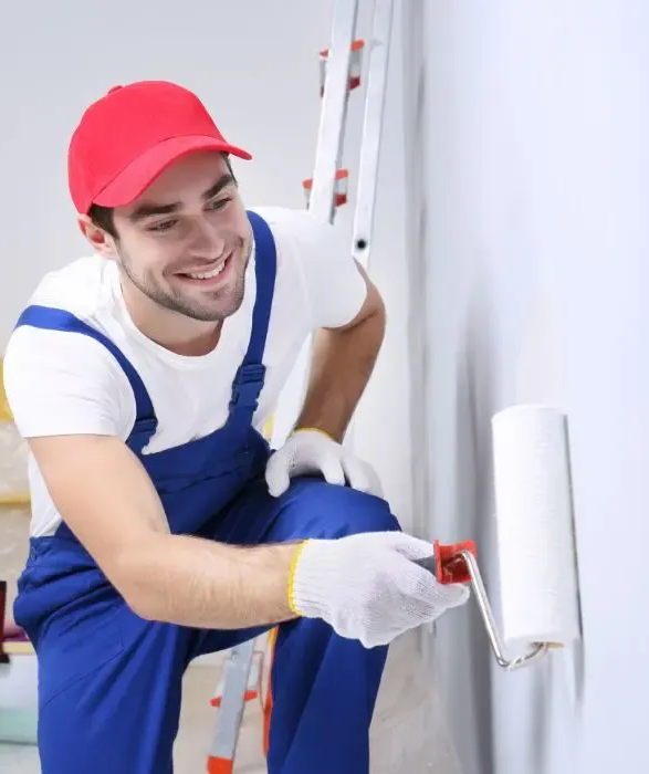 Painter in red cap and blue overalls applying white paint to a wall with a roller