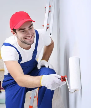 Painter in red cap and blue overalls applying white paint to a wall with a roller