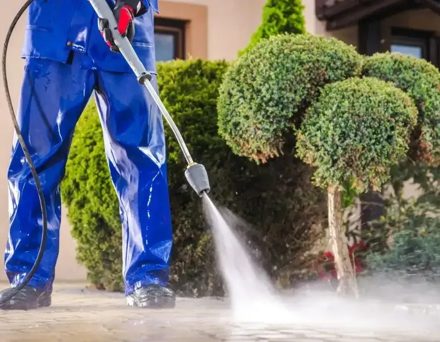 Person pressure washing a driveway beside trimmed bushes and a house