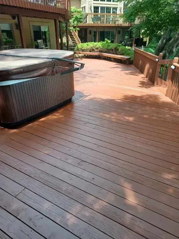 Wooden deck walkway beside a hot tub, bordered by railings and greenery, leading to buildings in the background