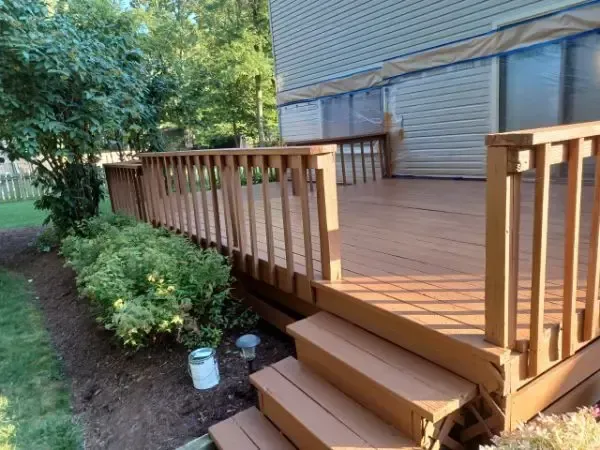 Wooden deck with steps beside a house, bordered by garden beds and trees.