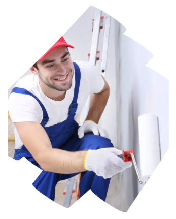 Painter in blue overalls and red cap smiling while applying white paint to a wall with a roller.