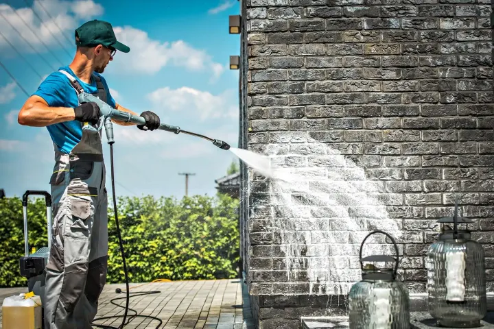 Worker pressure-washing a brick wall beside potted plants on a sunny patio