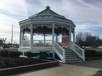 White gazebo with ornate trim and stairs in a park setting under an overcast sky