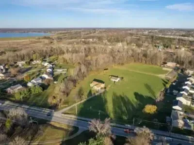 Aerial view of a grassy field beside a road, surrounded by trees and houses with water in the distance
