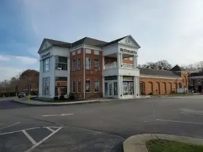 Two-story brick building with white columns beside an empty parking lot under a blue sky