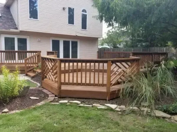 Backyard wooden deck with stairs and railings behind a beige house, surrounded by grass and landscaping.