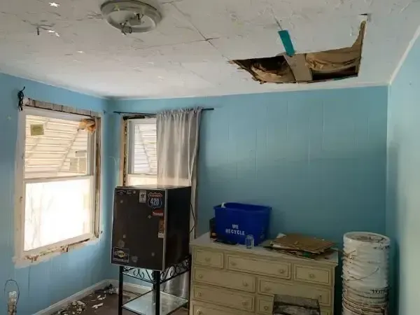 Damaged room with ceiling holes, peeling paint, two windows, and a dresser against blue walls
