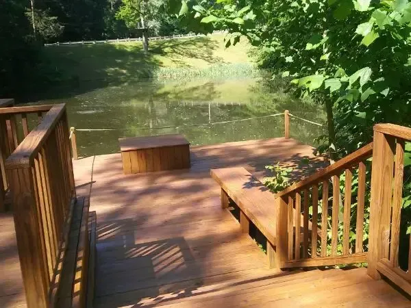 Wooden deck by a calm river, with railings, steps, and a bench in a shaded green setting