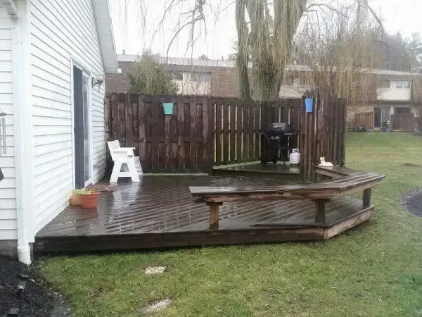 Small wooden backyard deck with a picnic table, white sign, and fenced yard on a rainy day