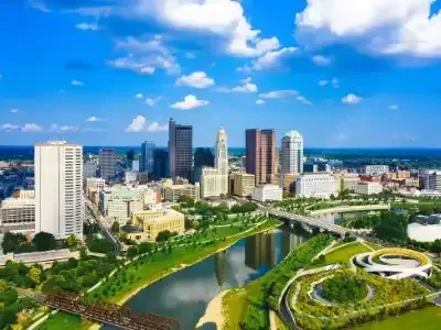 Aerial view of a city skyline beside a river and green park under a bright blue sky.