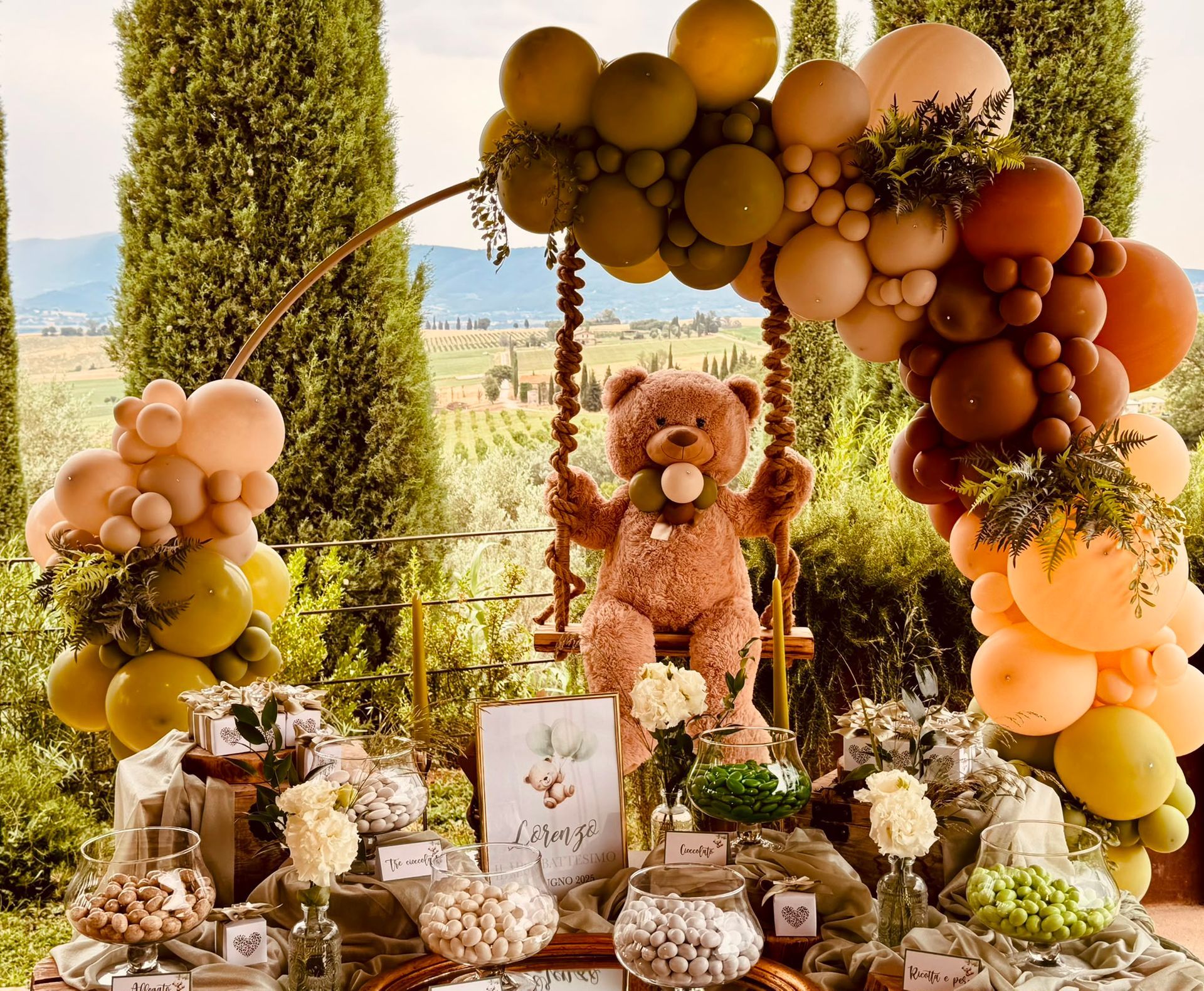 Allestimento per una festa a tema orsacchiotti con palloncini marroni, beige e color pesca, fiori e un centrotavola a forma di orsacchiotto sotto un arco all'aperto.