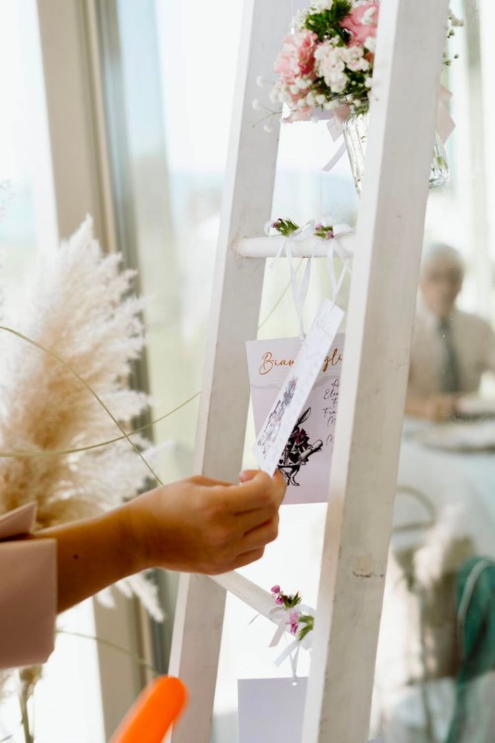 Una mano posiziona un biglietto su una scaletta bianca per gli auguri di matrimonio, decorata con fiori e nastri.