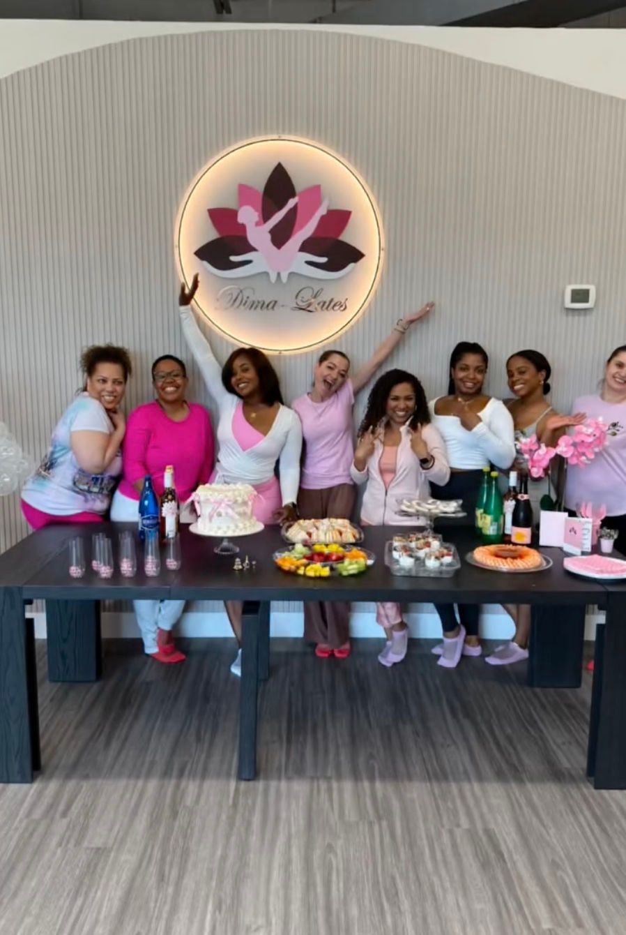 Group of people celebrating at a table with cake and drinks. Lotus logo in background. Mostly pink and white colors.
