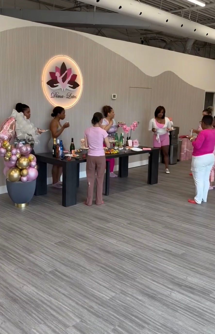 People at a party near a table with food, drinks, and decorations in a light-filled space.