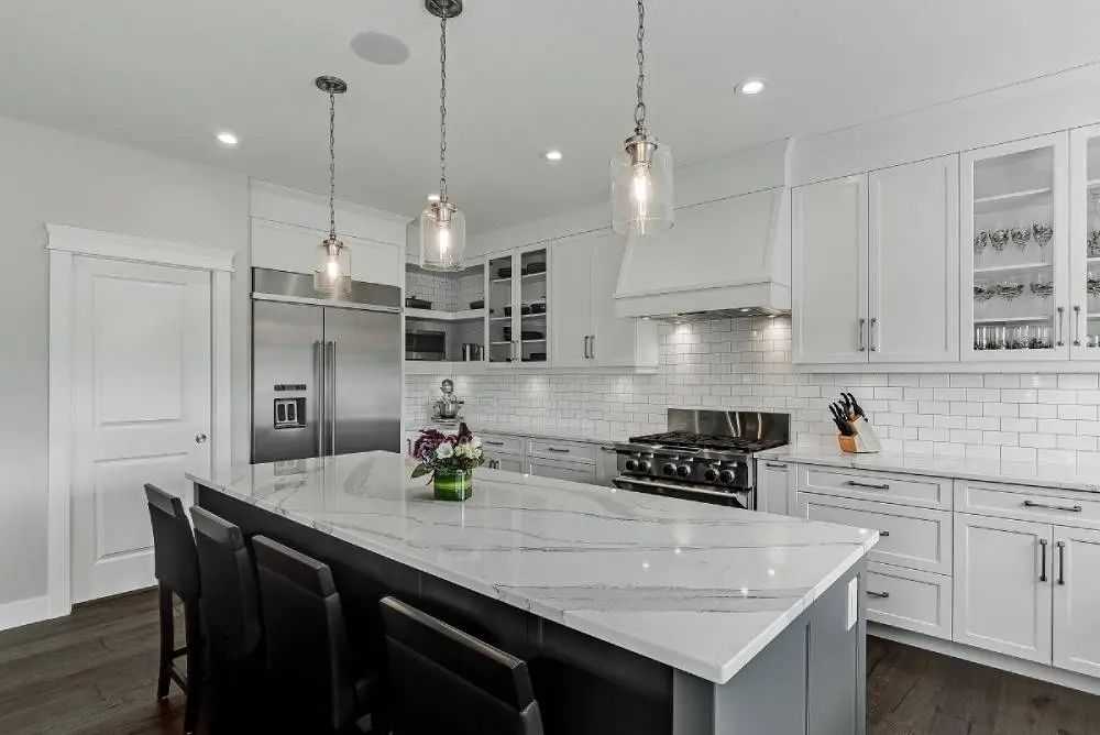 Modern white kitchen with island, marble countertop, stainless steel appliances, and pendant lights.