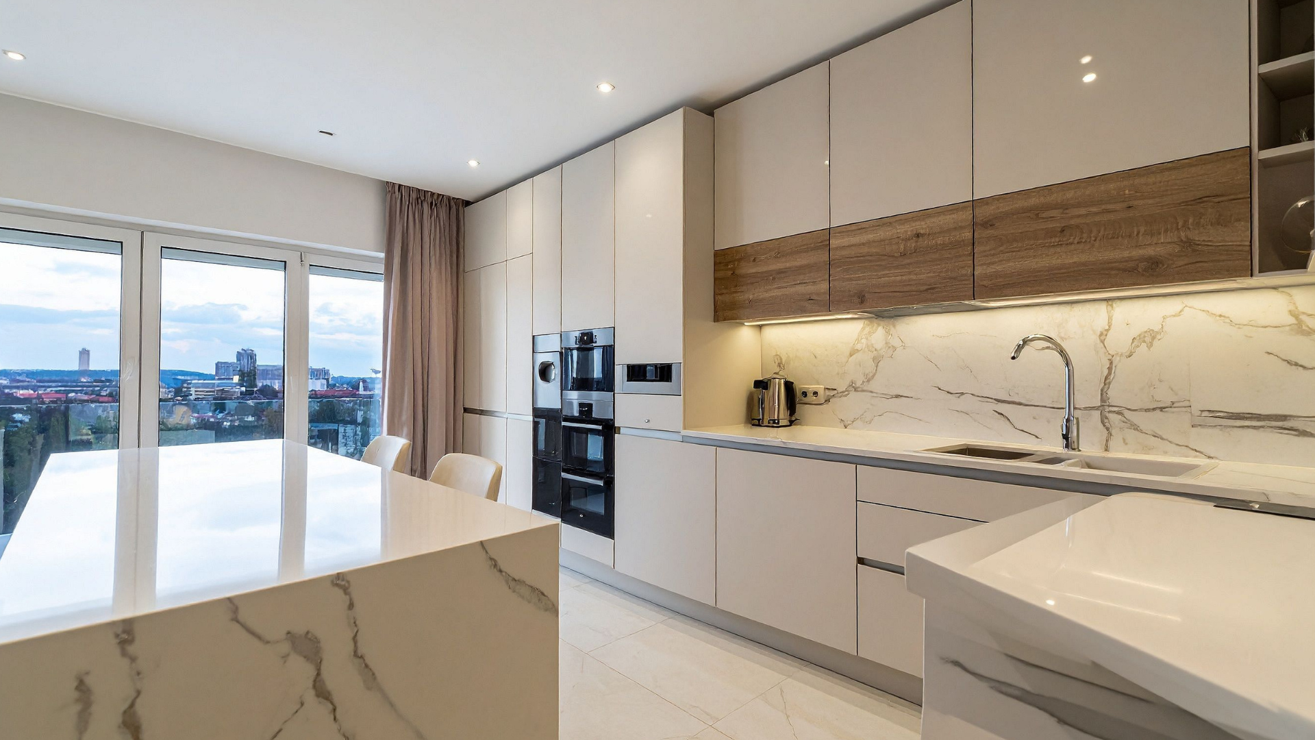 Modern kitchen with white cabinets, marble countertops, island, and large windows with a city view.