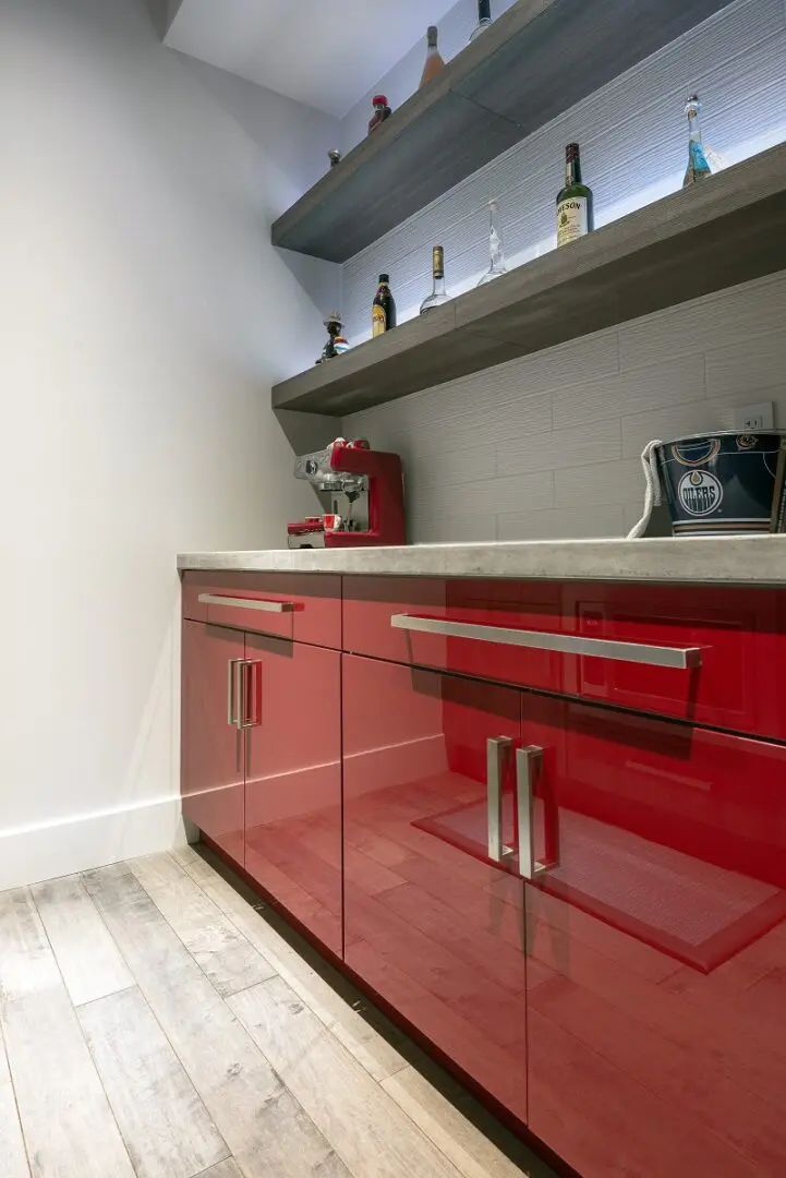 Red cabinets with silver handles under gray countertop. Bottles on shelves with under-shelf lighting.