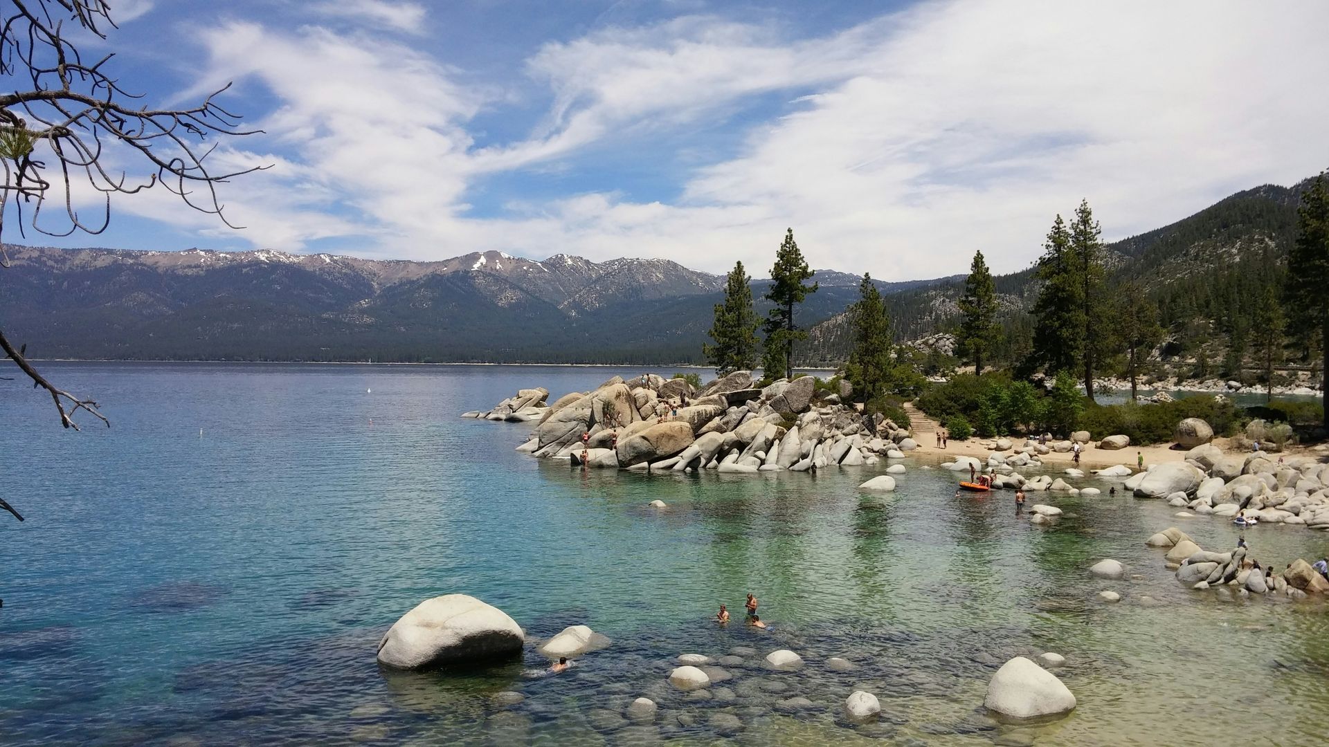 Lake Tahoe shoreline with clear water, rocks, trees, and distant mountains under a blue sky.
