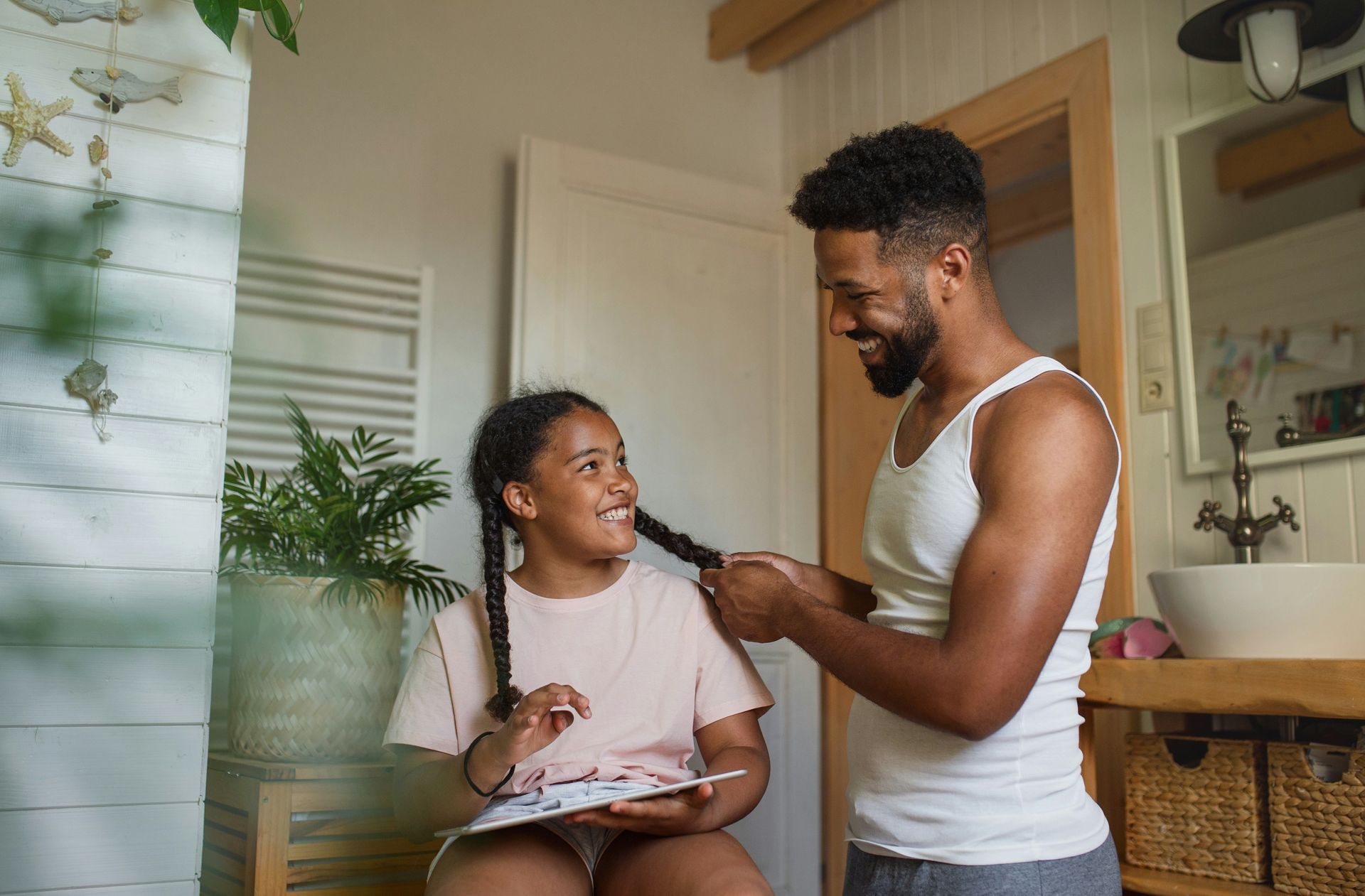 Dad braiding daughter's hair in bathroom, smiling at each other.