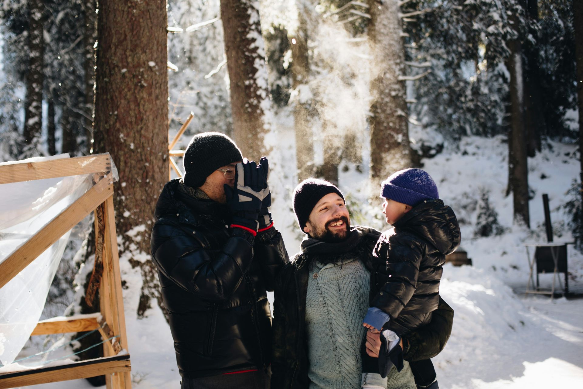 Two men and a child playing in snow, trees in the background. One man takes a photo, the other holds the child.