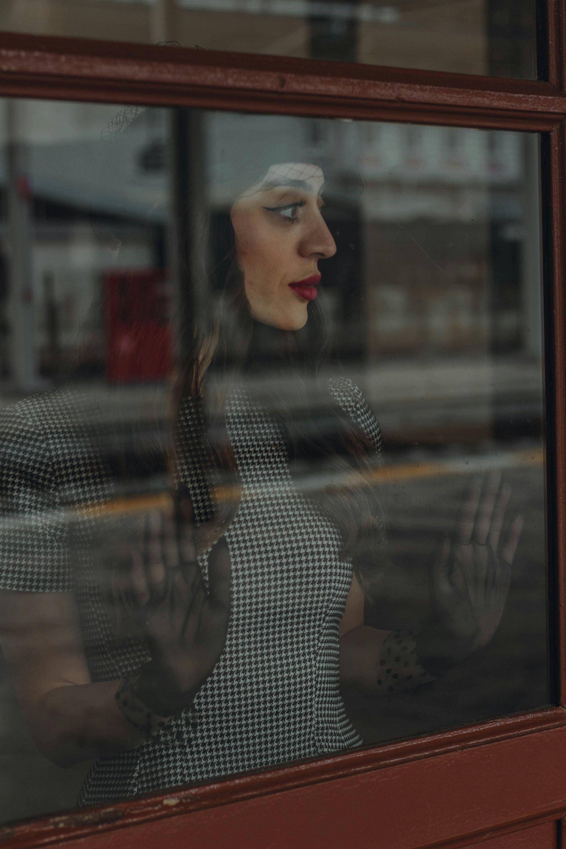 Woman with white makeup looks out a window, hands on the glass. Black and white dress, red lips.