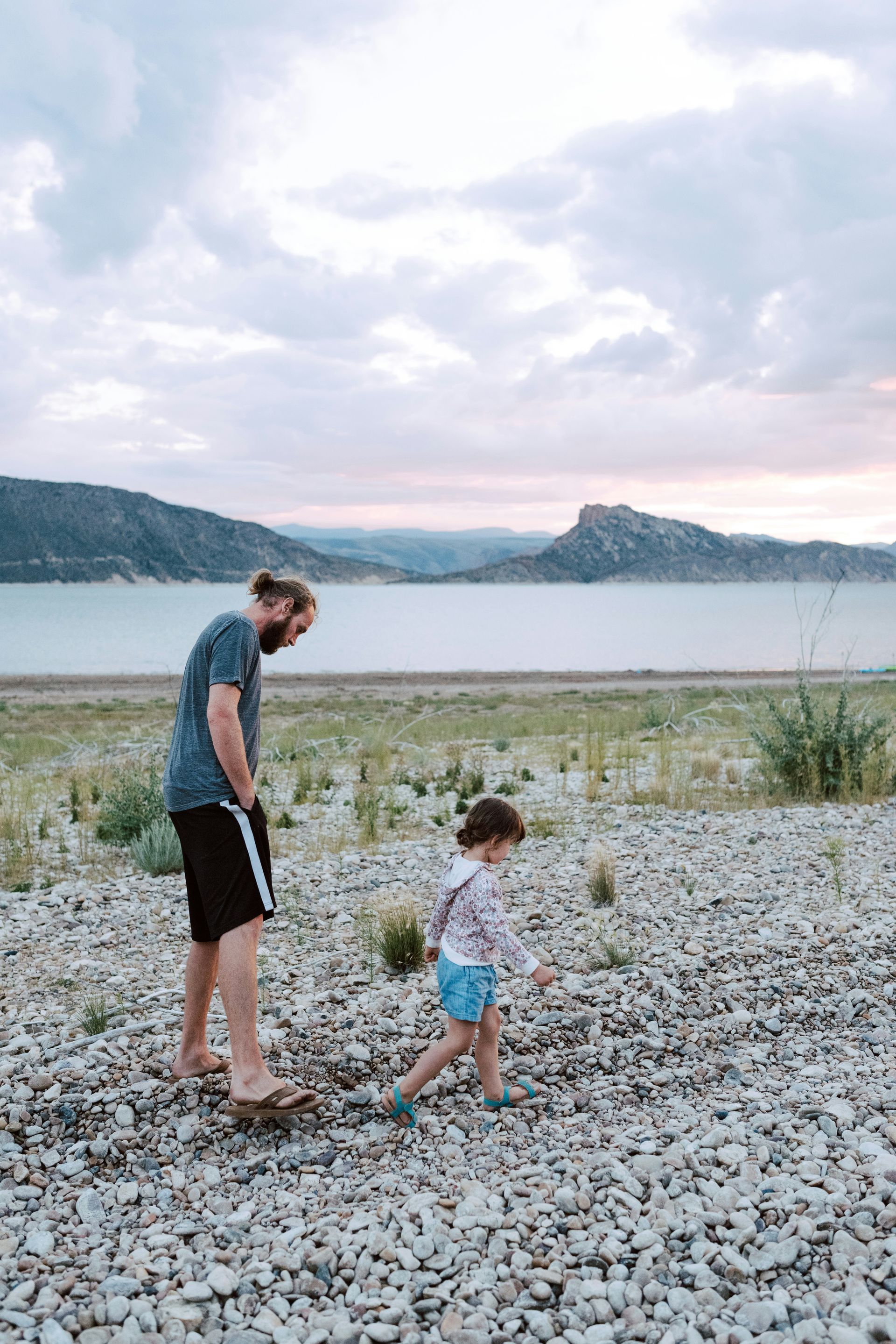 Father and child walking along a pebbled beach at sunset, with a lake and mountains in the background.