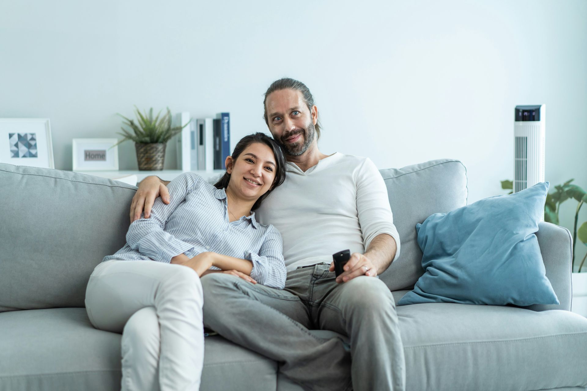 A smiling couple, embraced on a gray couch, holding a remote. Light-filled living room.