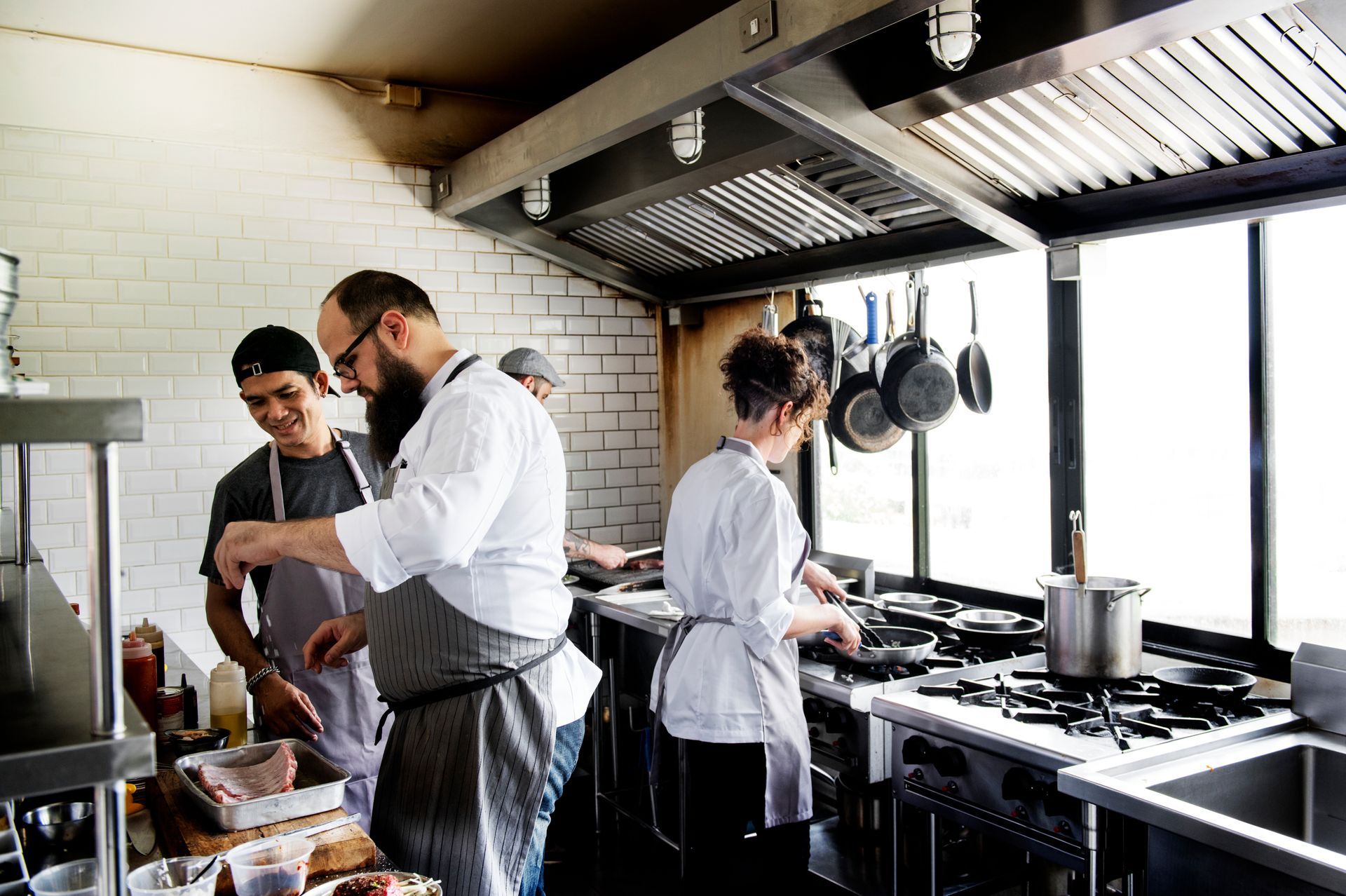 Chefs in a commercial kitchen, cooking food near a window, and an exhaust hood above a gas range.