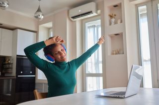Woman with headphones, arms raised, smiles while sitting at a table with a laptop in a well-lit room.