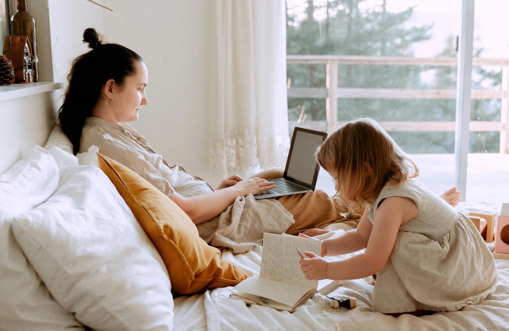 Woman working on laptop in bed; young child looks at a book. Light, airy room.