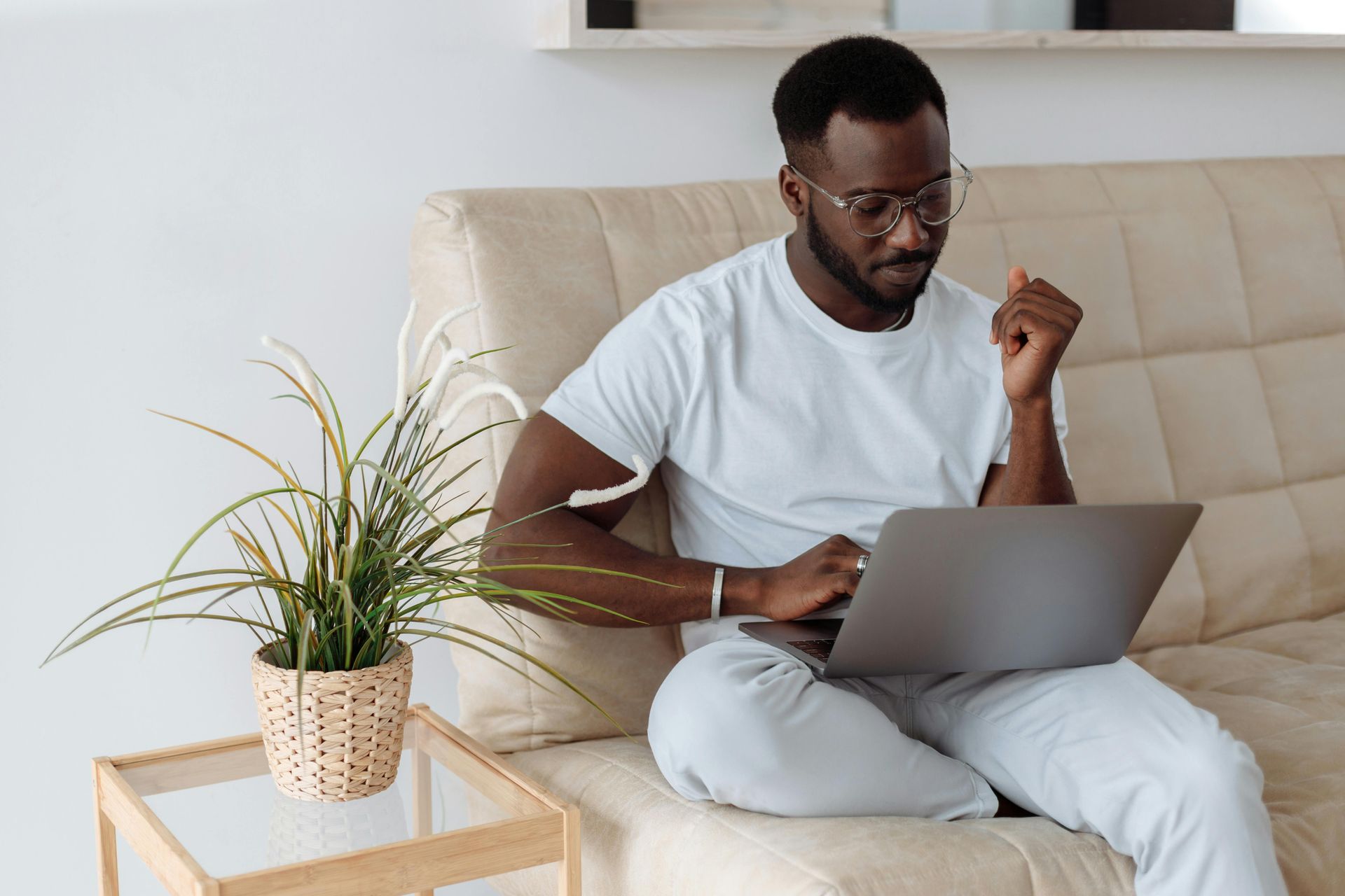 Man in glasses working on laptop on a couch next to a plant.