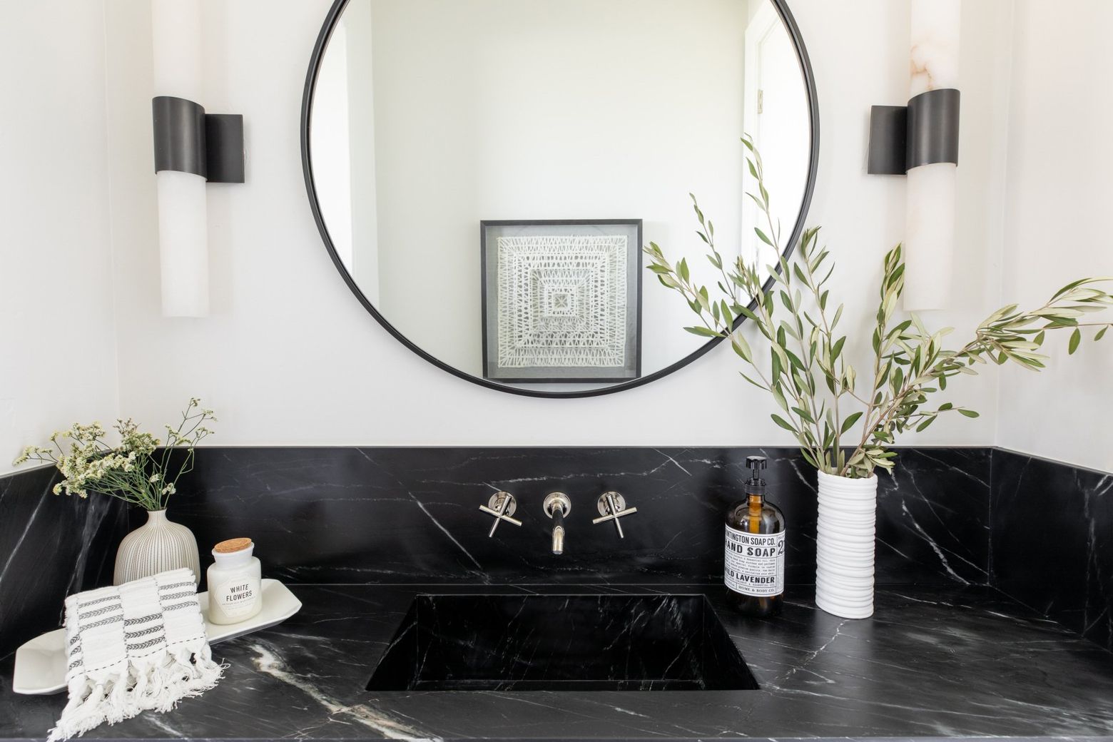 Modern bathroom vanity with black marble countertop, round mirror, wall sconces, and white vases with greenery