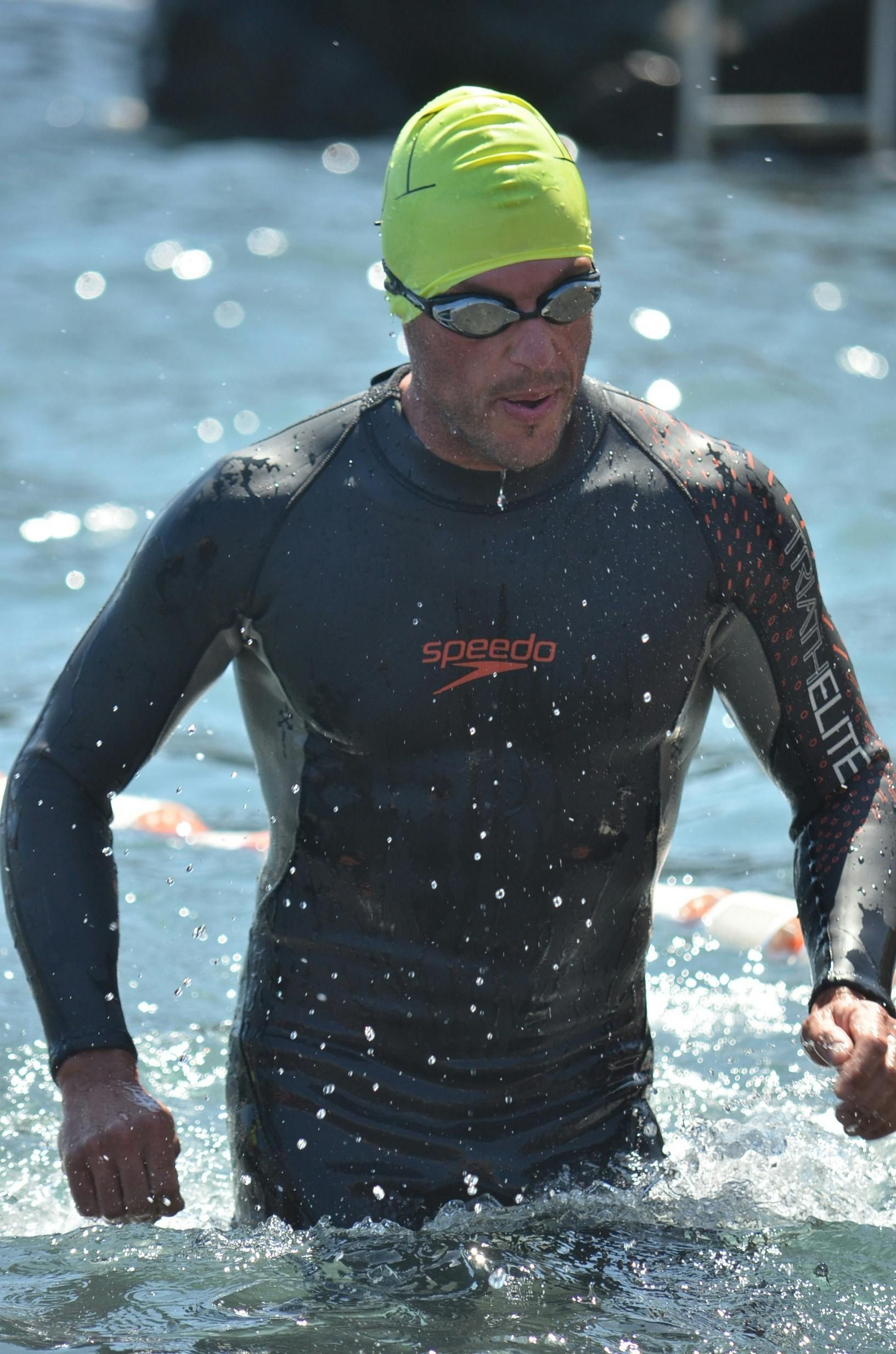 Een man in een wetsuit en badmuts komt uit het water na het zwemmen. Hij draagt een duikbril; het water spat om hem heen.