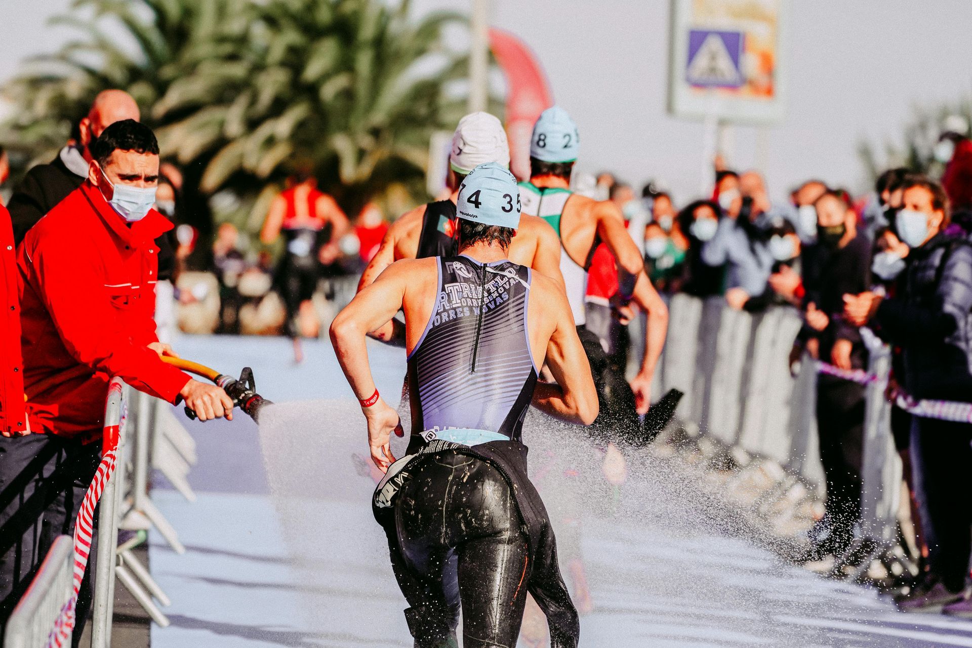 Triatleten komen uit het water en rennen richting de wisselzone. Toeschouwers met maskers kijken toe en iemand sproeit water.
