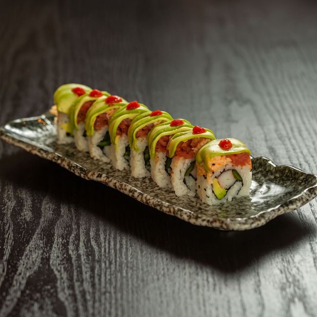 Sushi roll topped with avocado and red sauce on a rectangular plate