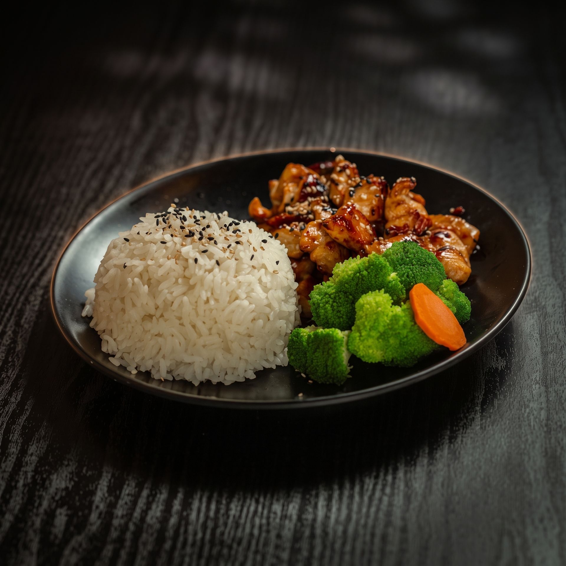 Black plate with rice, glazed chicken, broccoli, and carrots on a dark table