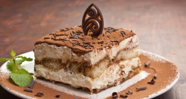 Tiramisu dessert square on a white plate, topped with cocoa and chocolate decoration, with mint garnish.