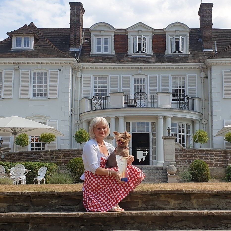 A middle-aged woman perched on stone steps before a grand white hotel, her dog and an award held within her arms.