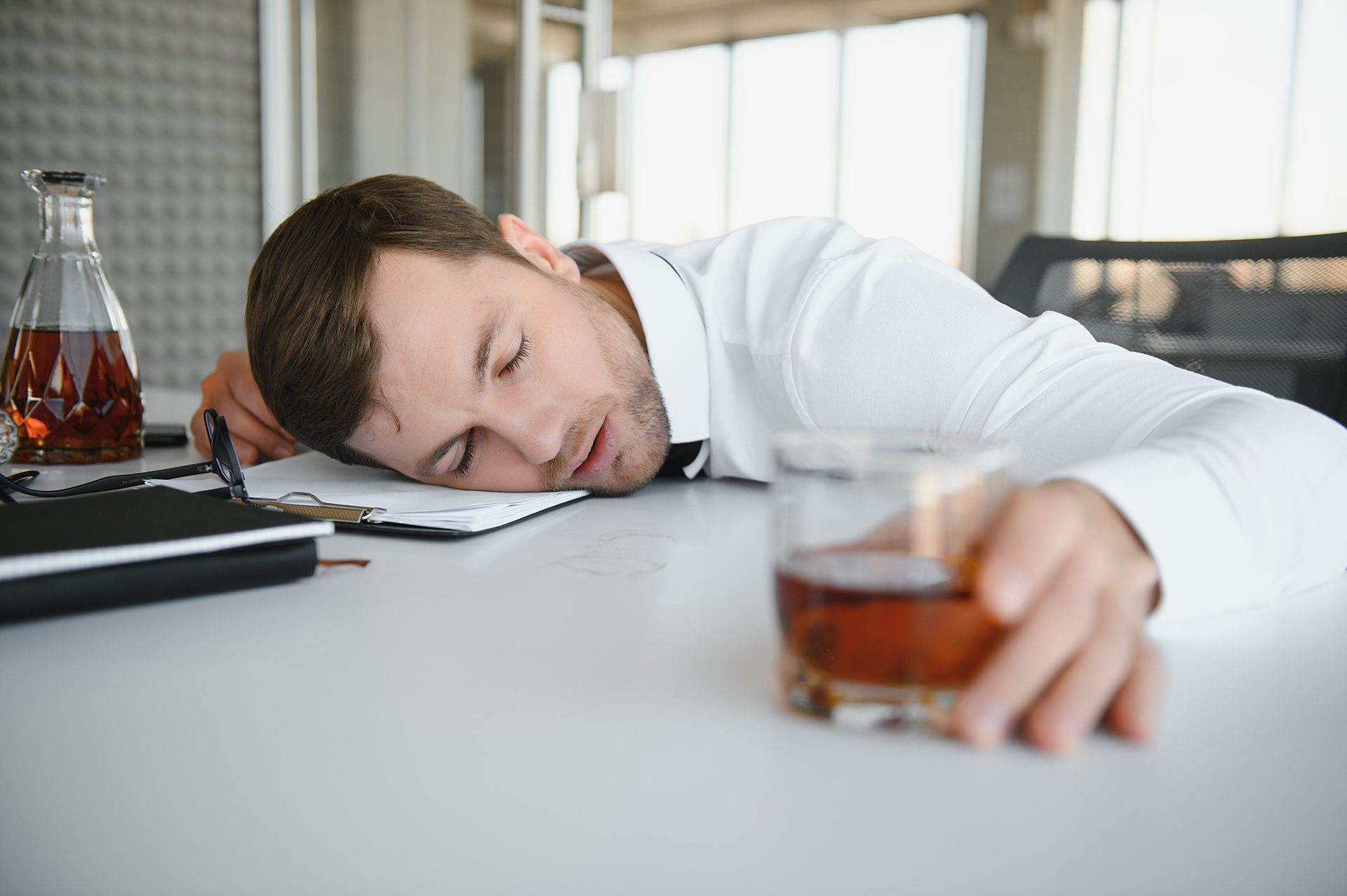 Man asleep at a desk, head on papers, with a glass of whiskey in front of him.
