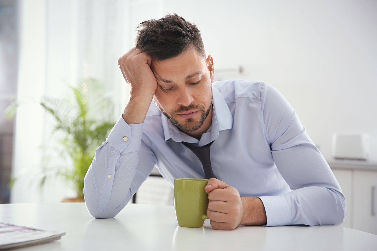 A tired person in a dress shirt and tie leans on a table holding a green mug in a bright room.